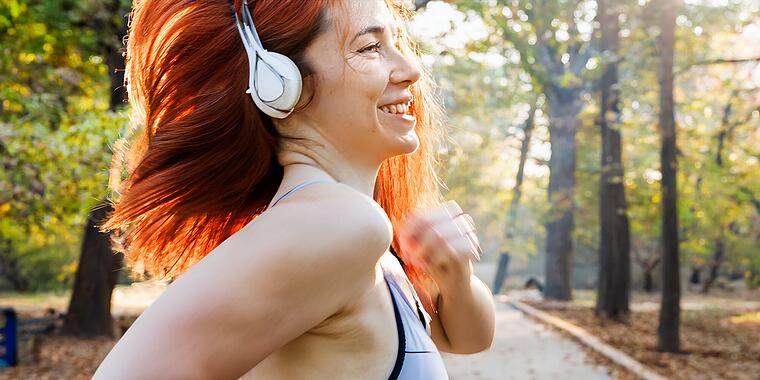 PLUS-Audio-Visual Eine junge rothaarige Frau joggt auf einem leicht verschwommenen Parkweg im sanften Morgenlicht. Sie trägt moderne Sportkleidung, darunter Laufleggings und ein atmungsaktives Top, sowie große Over-Ear-Kopfhörer auf dem Kopf. Die Frau ist im Vordergrund leicht seitlich von vorne zu sehen, wirkt fokussiert und entspannt, mit einem dezenten Lächeln. Im Hintergrund ist ein grüner Stadtpark mit Bäumen zu erkennen, mit einem sanften Bokeh-Effekt und warmen natürlichen Farben. Das Bild zeigt eine dynamische Bewegung im Laufmoment, realistische Licht- und Schatteneffekte, hohe Detailgenauigkeit und natürliche Hauttöne.