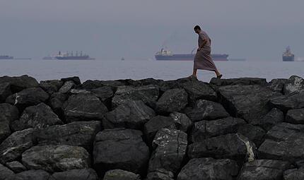Ein Mann geht auf den Felsen am Ufer spazieren, während Öltanker und Frachtschiffe in der Straße von Hormus aufgereiht sind. Ein Mann geht auf den Felsen am Ufer spazieren, während Öltanker und Frachtschiffe in der Straße von Hormus aufgereiht sind.
